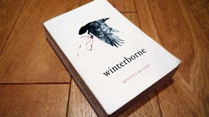 Winterborne book on floor featuring a crow flying on a white background carrying a red ribbon